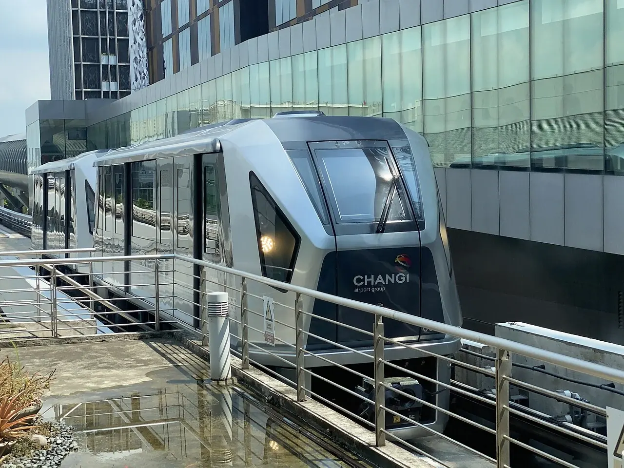 A silver airport train has a pointed dark front window and lit headlights.