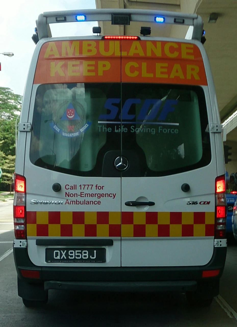 A white ambulance is seen from behind, with bright warning lights and checked markings.
