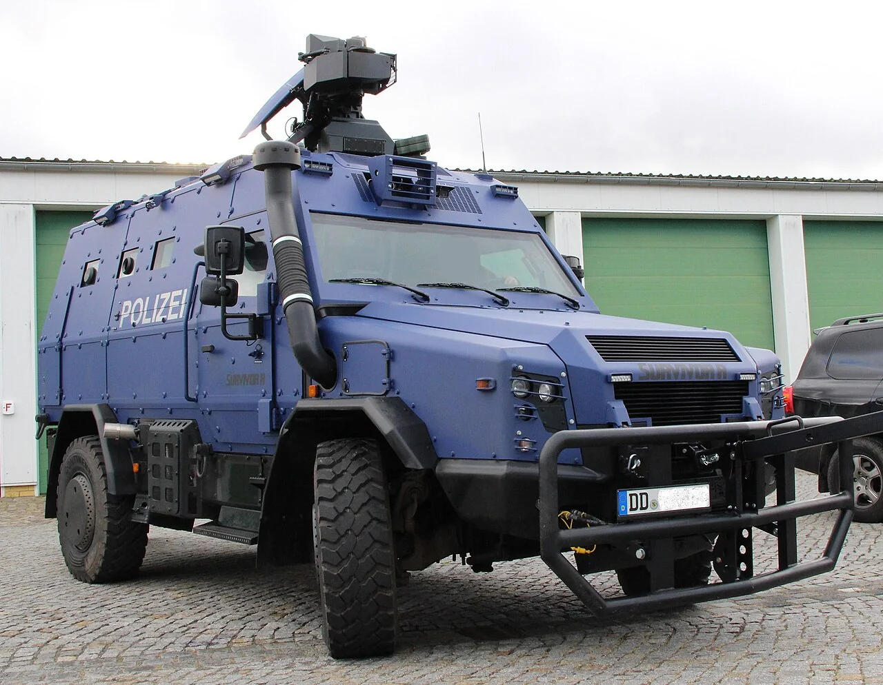 A blue armoured police vehicle with large tyres and a roof-mounted camera is parked outside a building.
