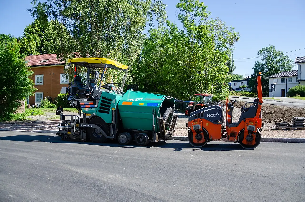 A green asphalt paver and an orange road roller stand beside a road construction site.