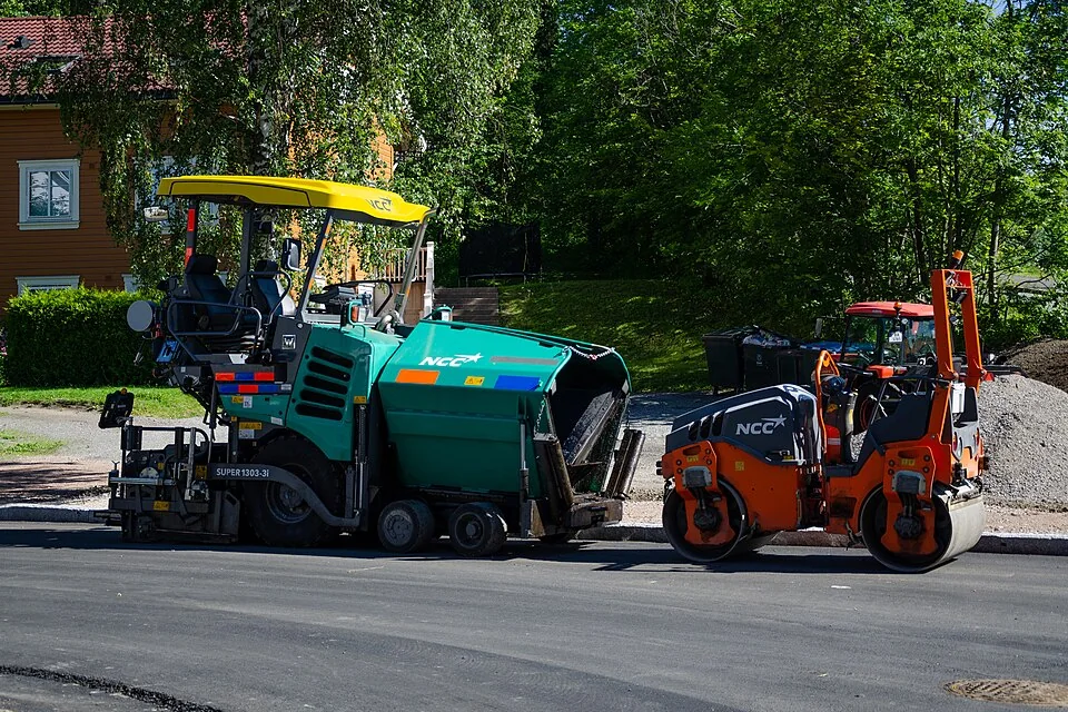 A green paving machine stands beside a small orange roller on a dark road.