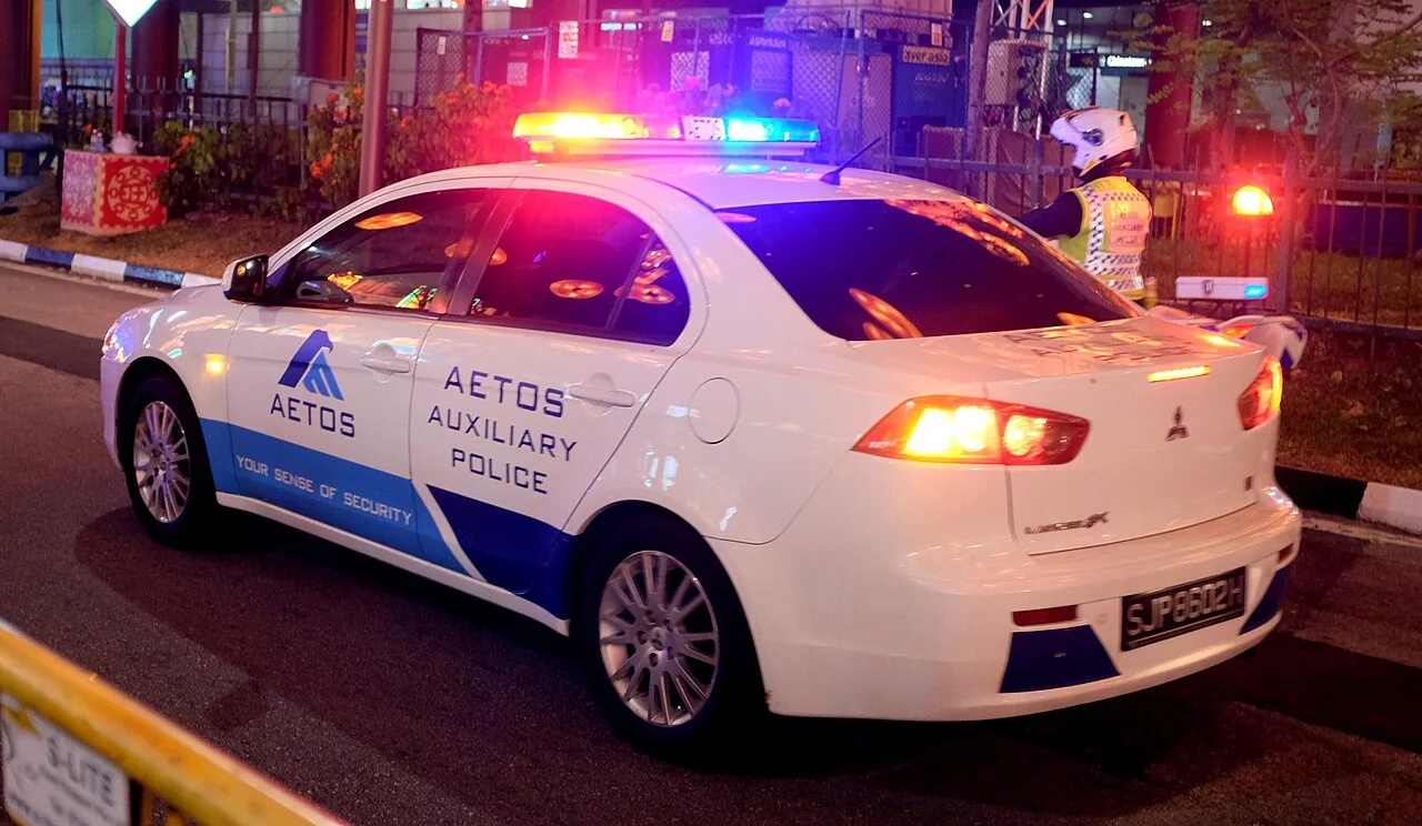 A white AETOS Auxiliary Police car has blue markings and glowing roof lights at night.