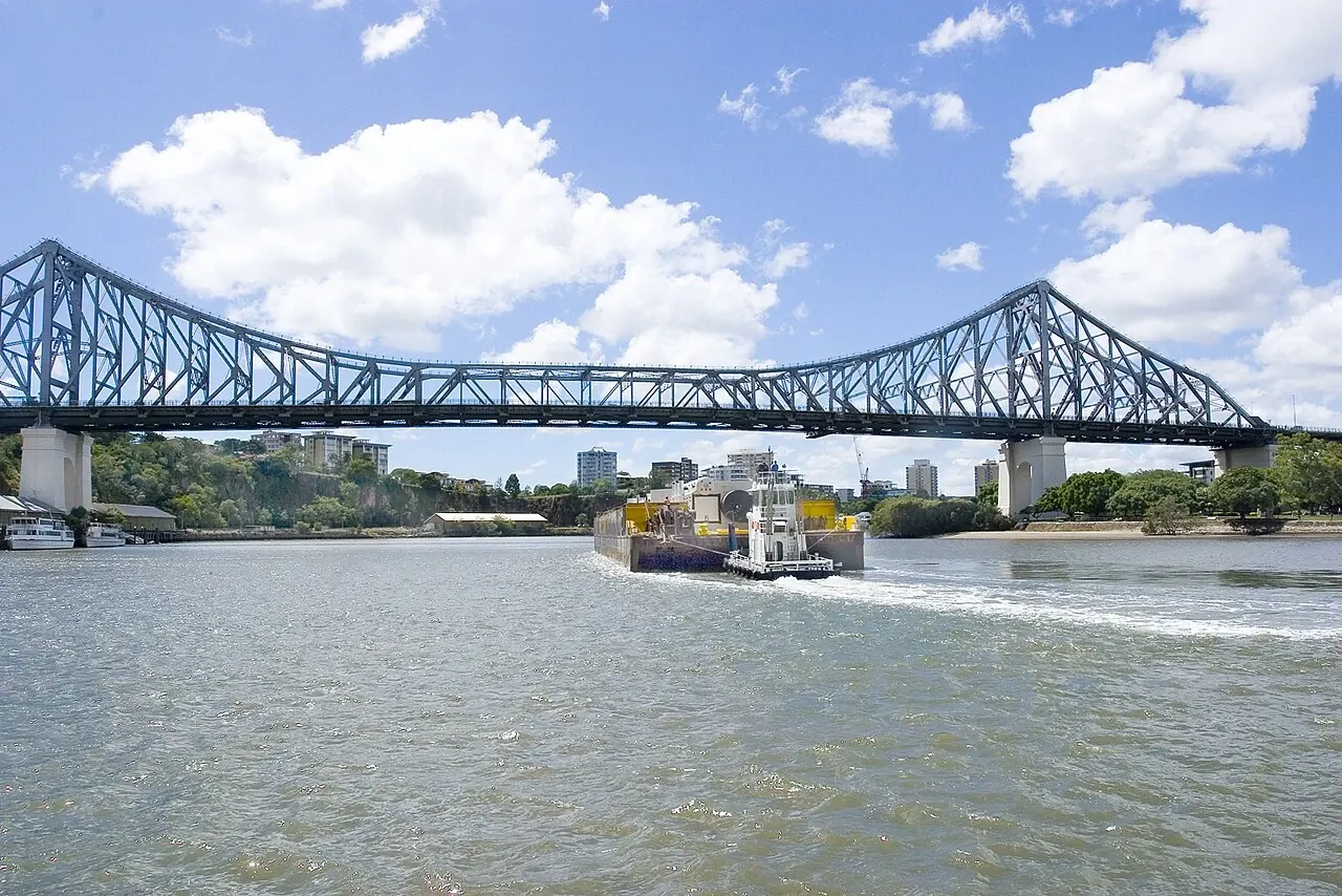 A tug pushes a loaded barge towards a large steel bridge.