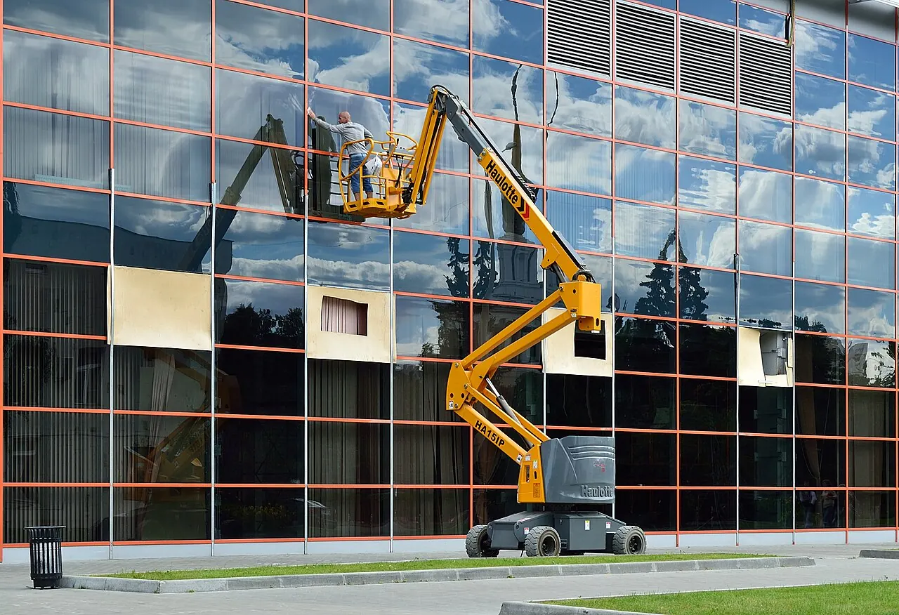 An articulated boom lift raises a window cleaner beside a glass building.