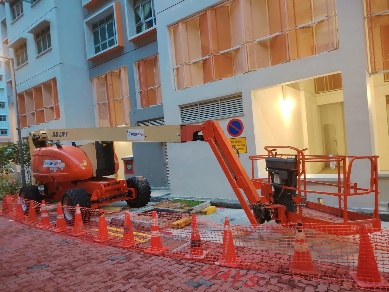 A boom lift with its long arm lowered stands beside a residential building, behind cones and safety mesh.
