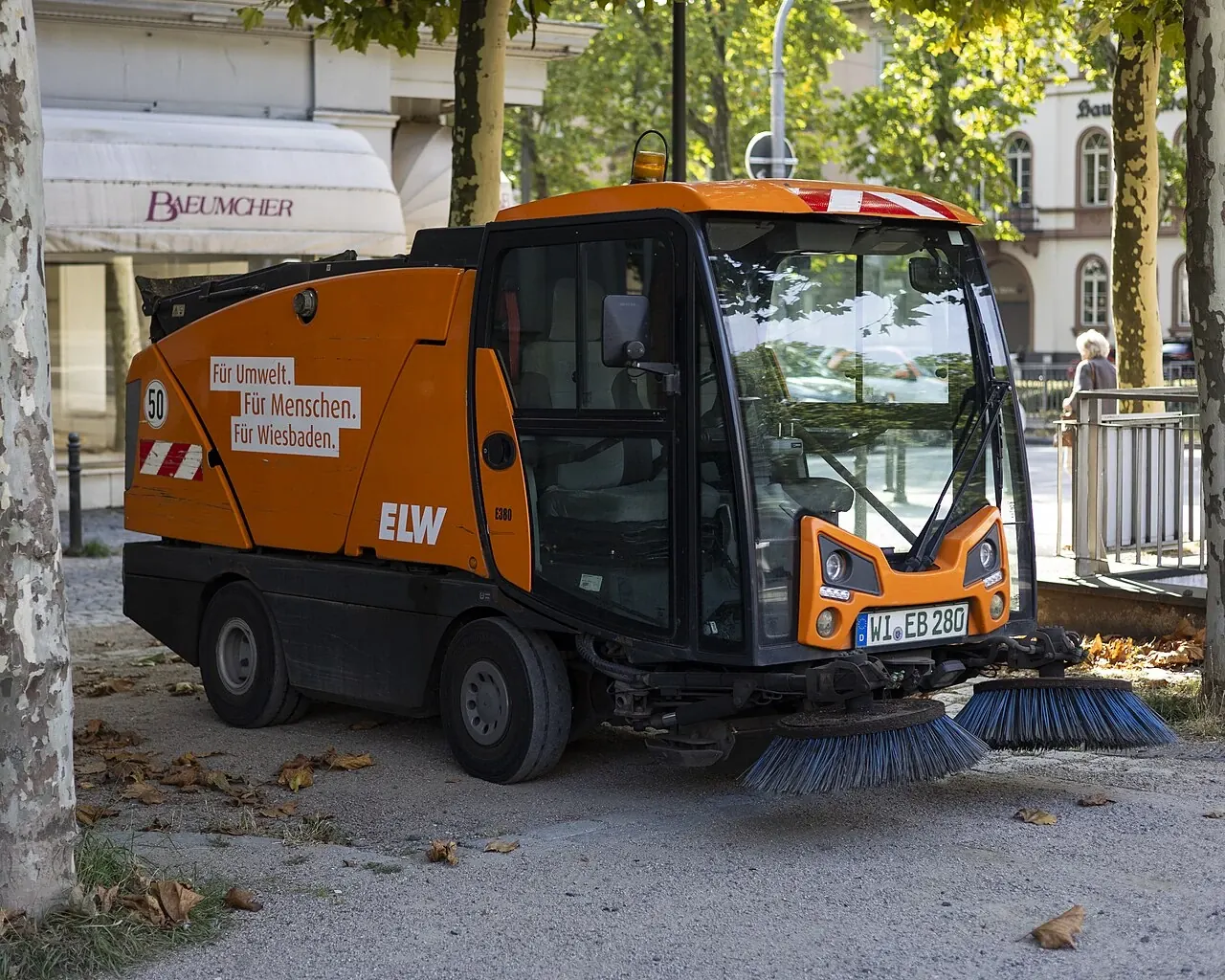 An orange compact sweeper has two round brushes beneath its front windscreen.