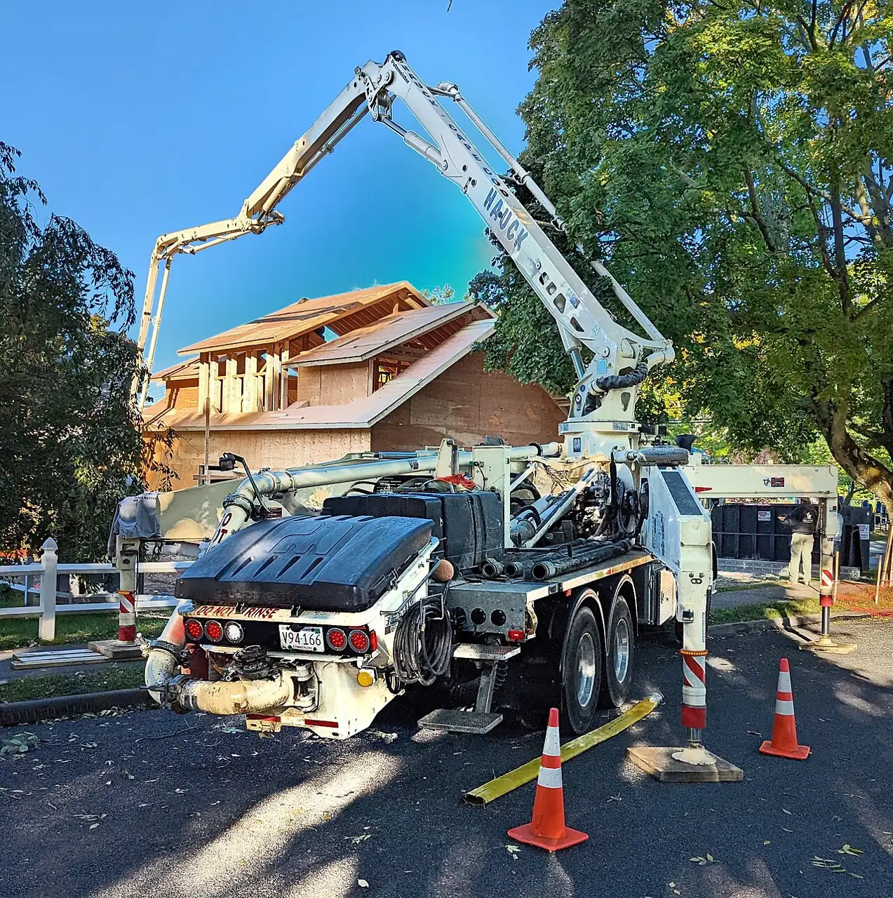 A concrete pump truck has a raised white boom beside orange traffic cones and trees.