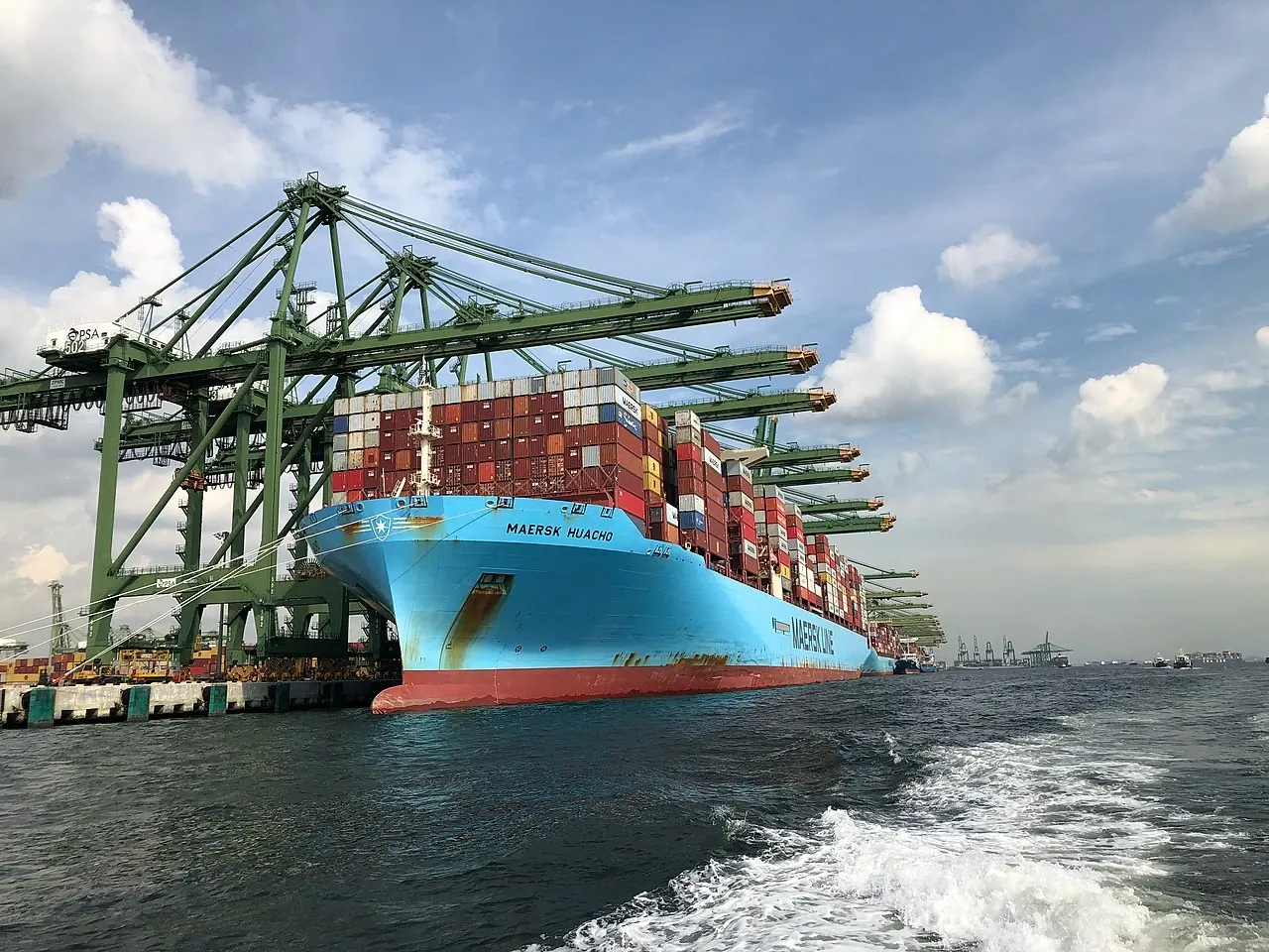 A blue container ship sits beside large green cranes, with colourful containers stacked on deck.