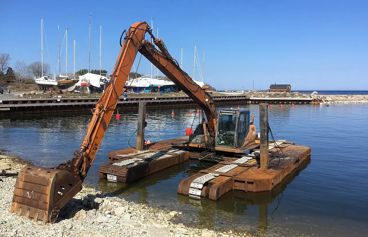 An orange excavator with broad pontoon tracks rests at the edge of the harbour.