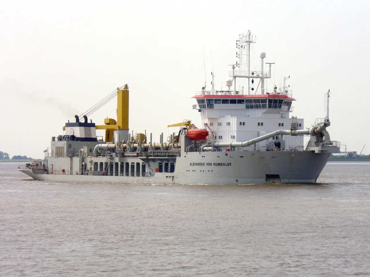 A grey dredger has a tall white bridge and yellow equipment along its deck.