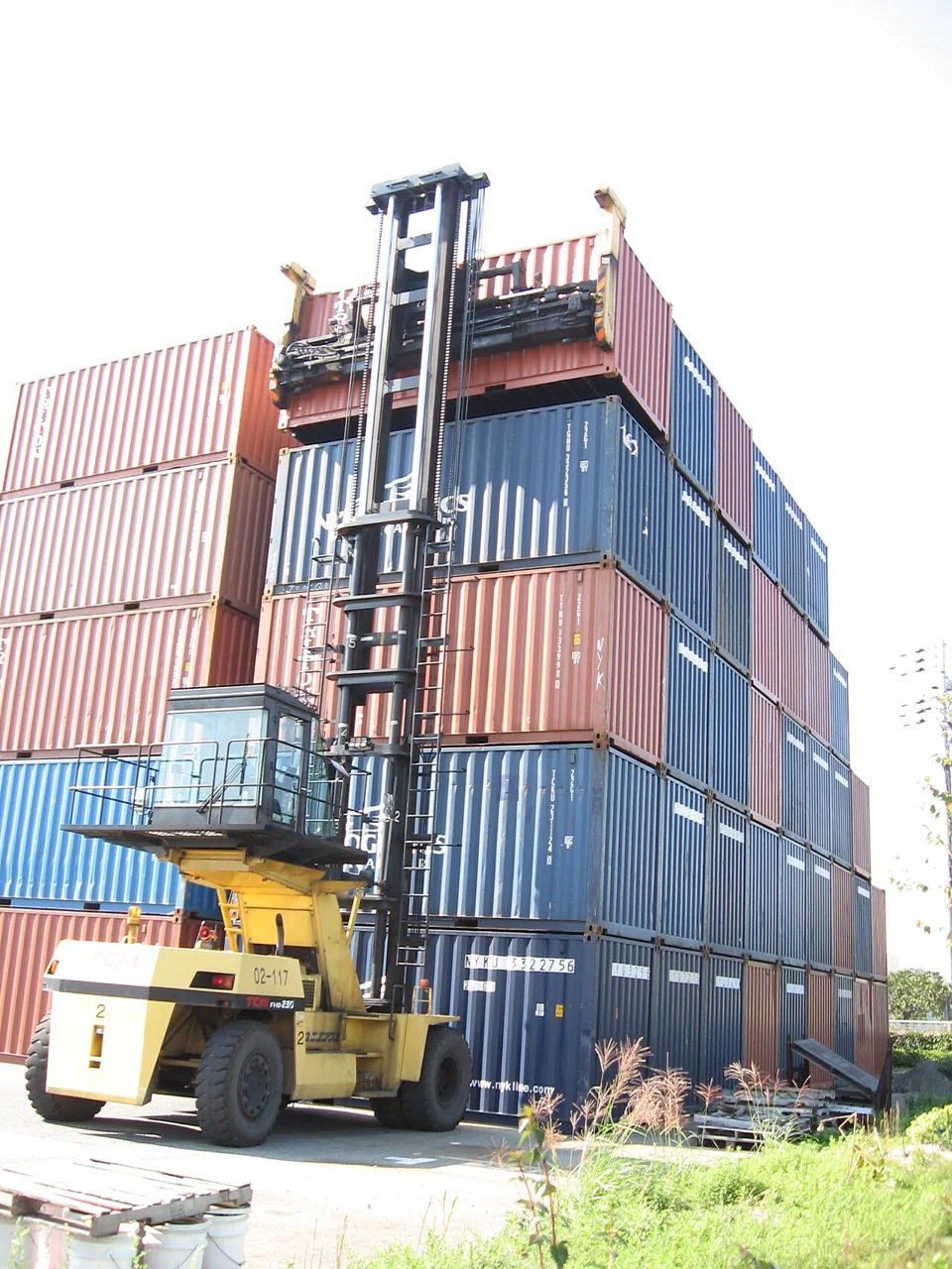 A yellow empty-container handler raises its tall mast beside a stack of red and blue containers.