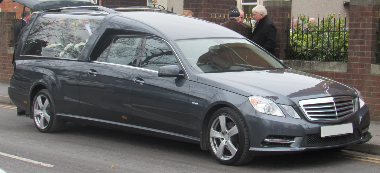 A long grey hearse has a raised rear roof and large windows, with flowers visible inside.