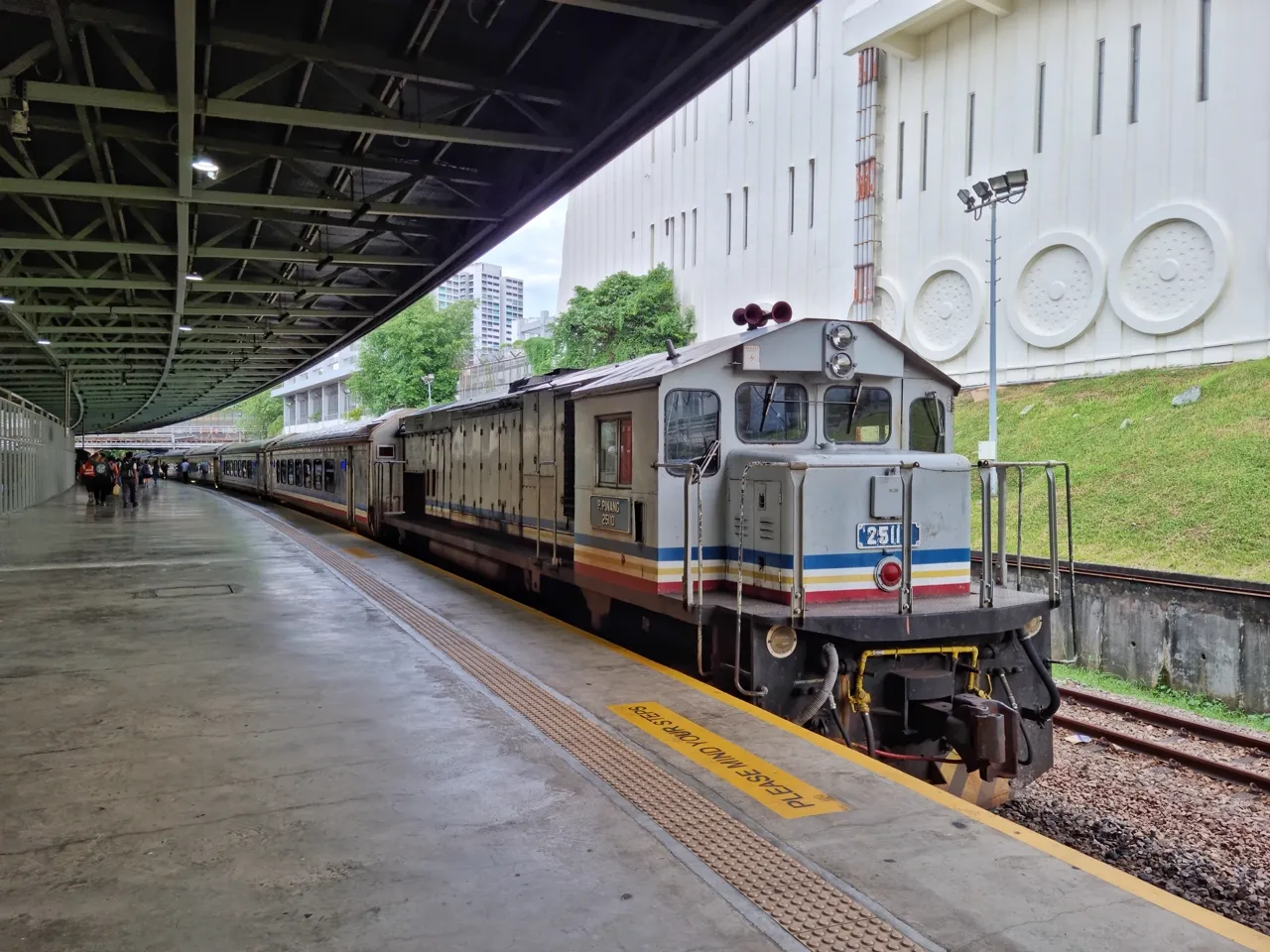 A grey diesel locomotive pulls coaches beside a covered railway platform.