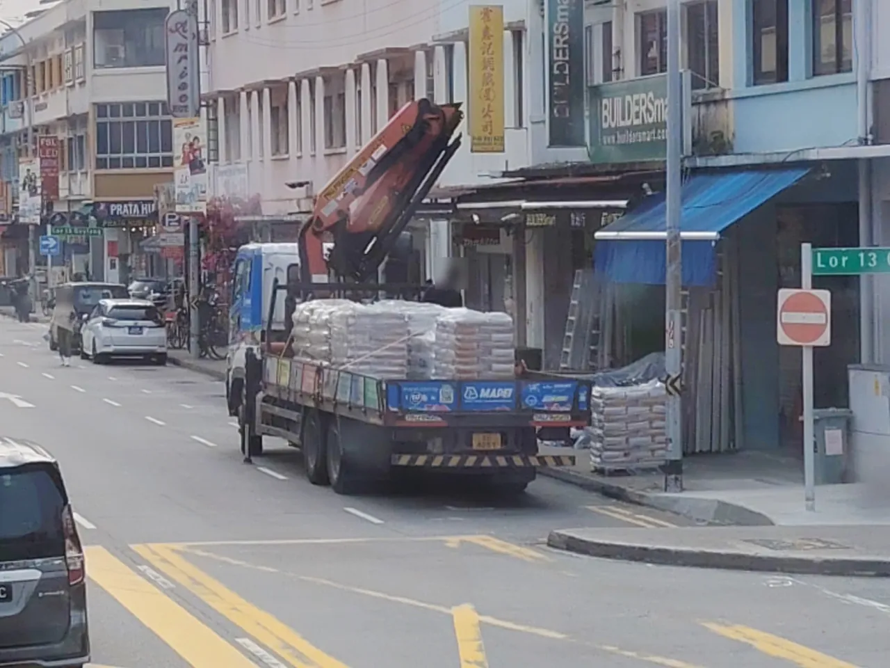 A blue lorry carries stacks of white bags beneath its raised orange crane arm.