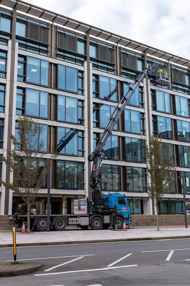 A blue lorry has a tall crane arm raised beside a glass-fronted building.