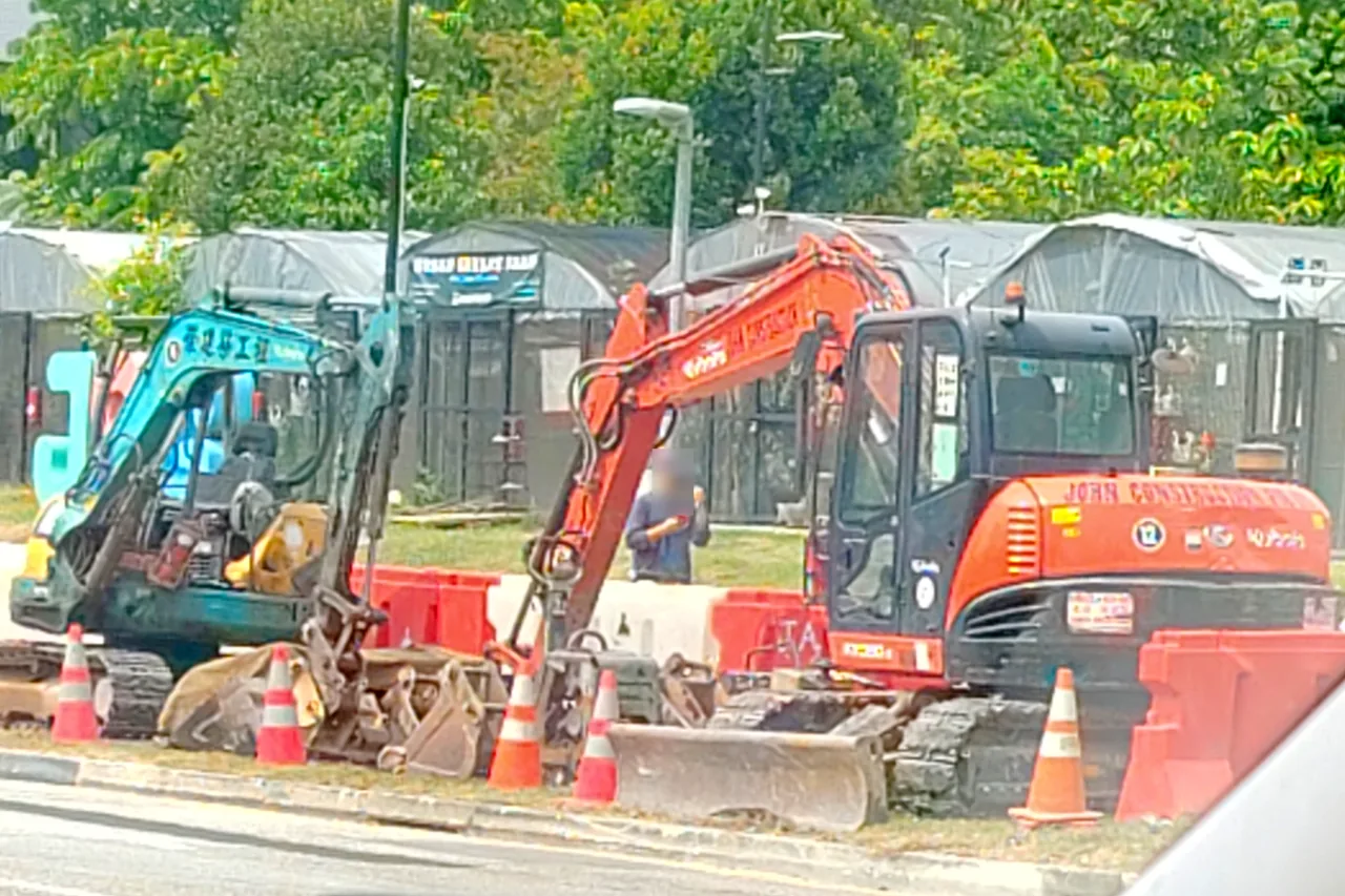 Mini excavator — an orange mini excavator and a blue-green one parked at a roadside works site with cones and barriers, Photographed in Singapore