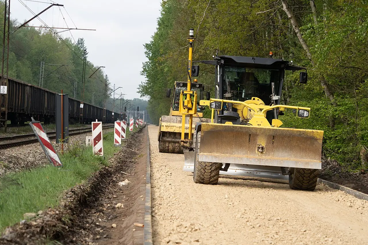 A yellow motor grader uses its long blade on a roadwork site beside railway tracks.