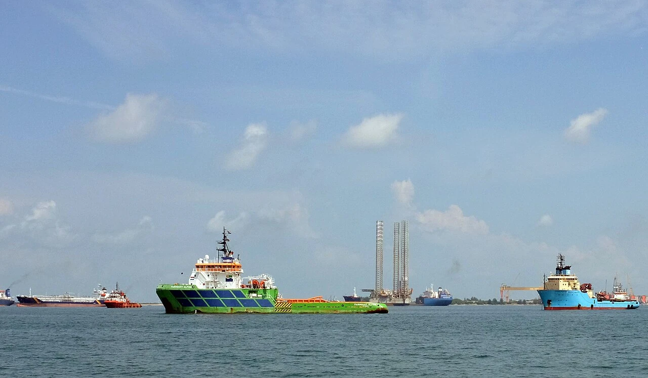 A green support ship floats between other vessels on calm blue water.