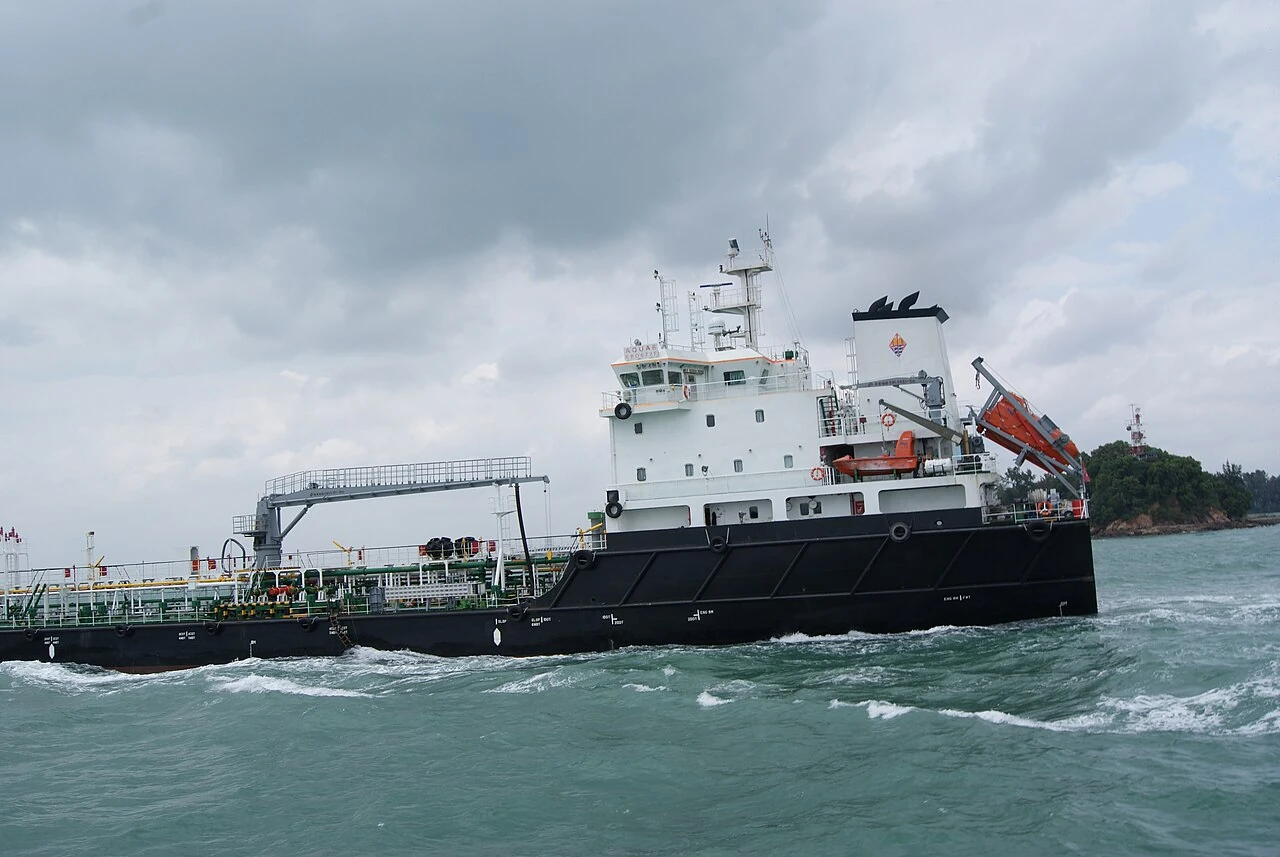 A black tanker has a tall white bridge above its pipe-covered deck.