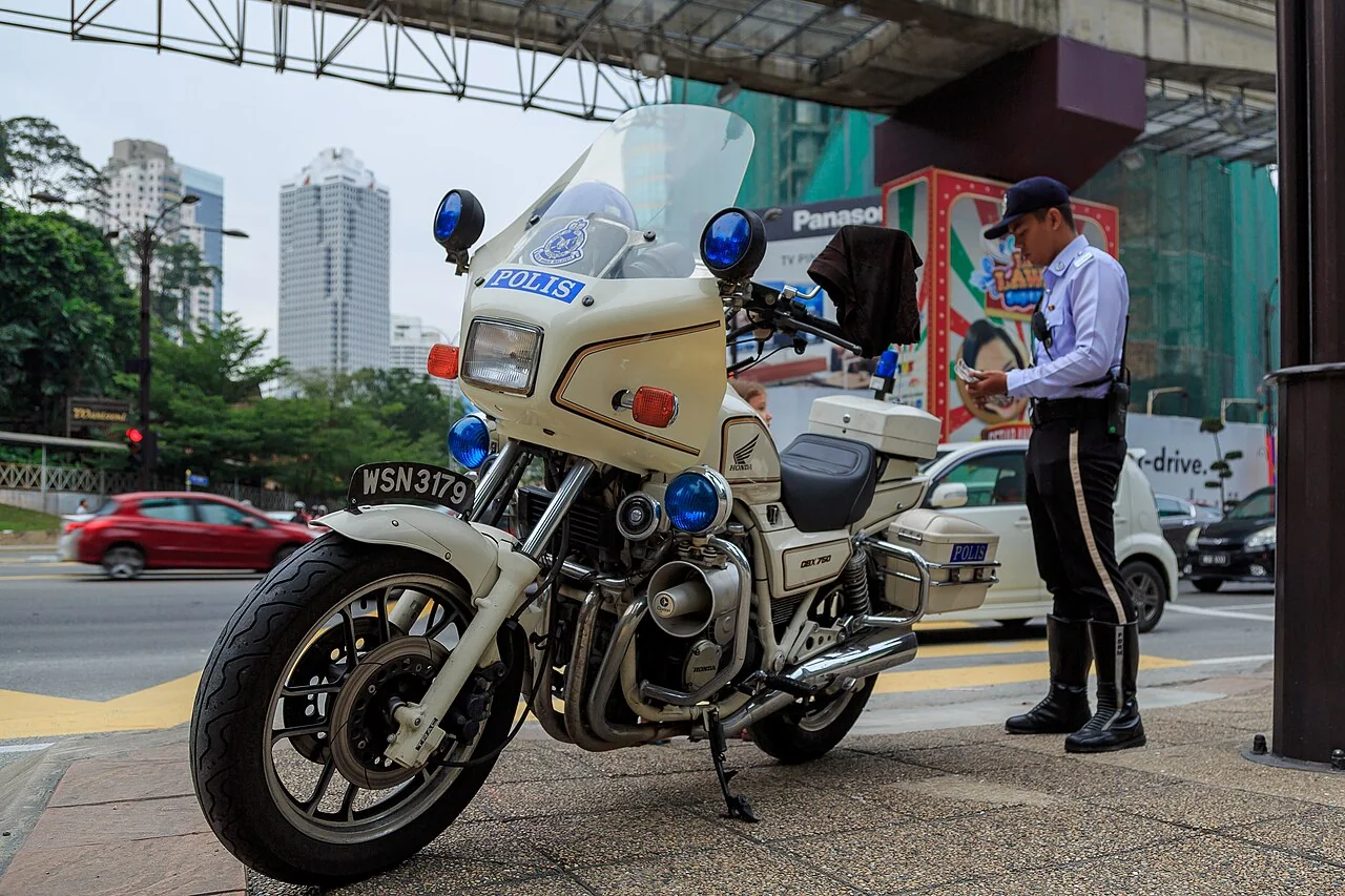 A traffic police officer in uniform stands beside a white Honda police motorcycle marked 'POLIS' at a city intersection.