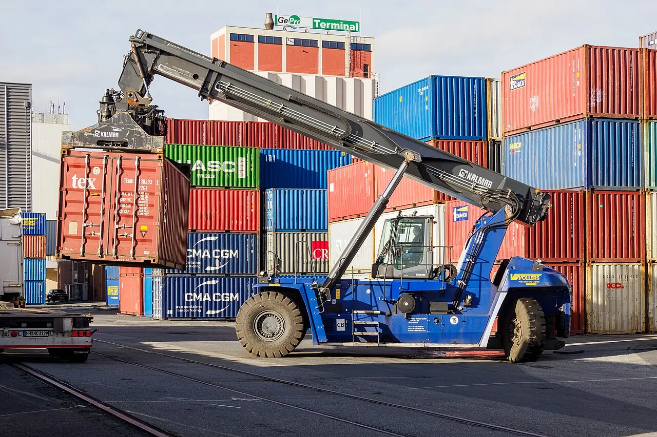 A reach stacker raises its long boom beside stacks of containers.