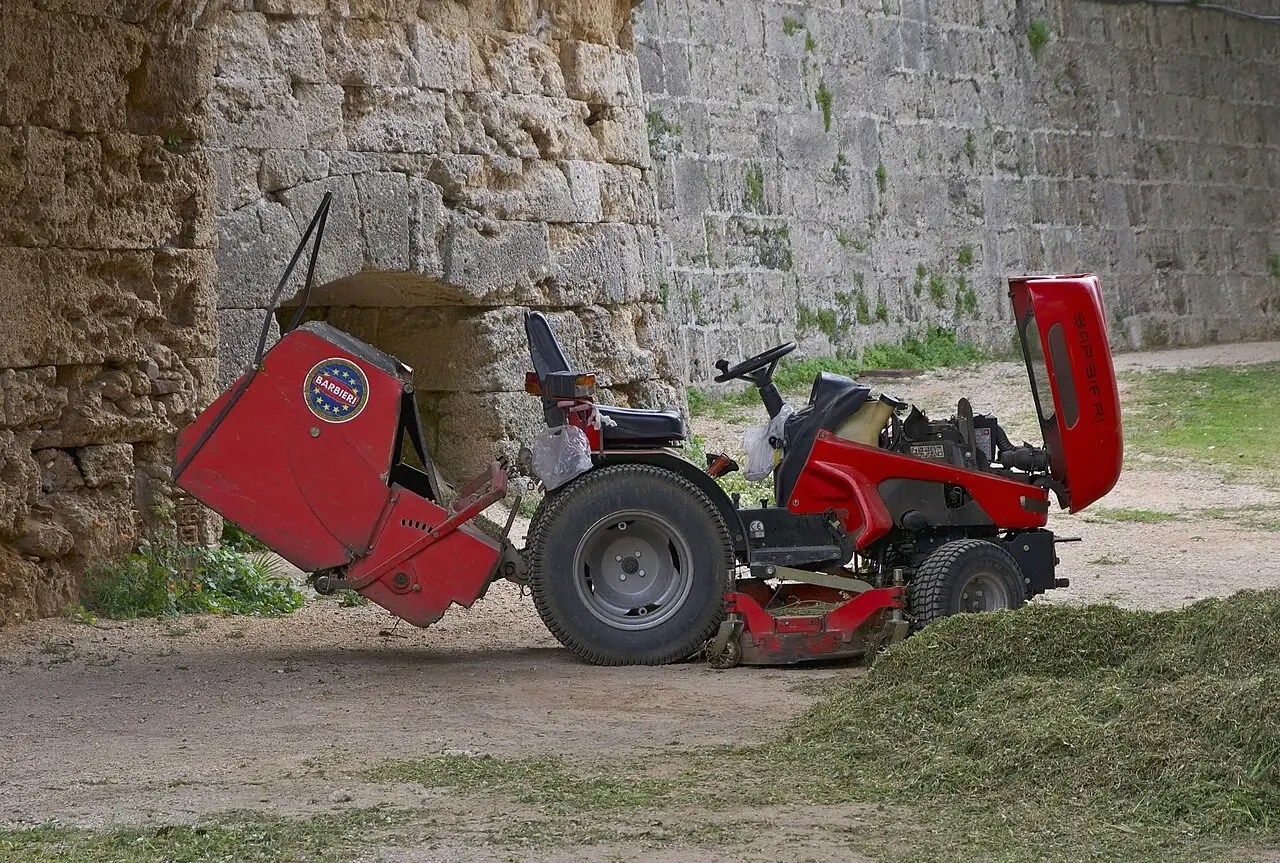A red ride-on mower has its rear grass box tipped up beside a pile of cut grass.