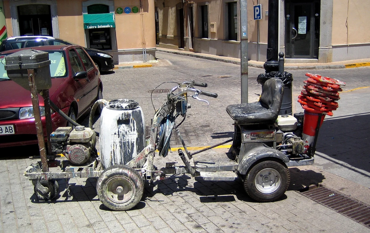 A road-marking machine has a paint-splashed tank at the front and a seat beside stacked orange cones at the back.