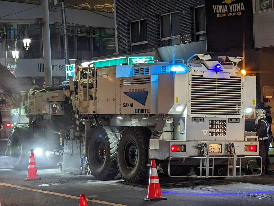 A large white road machine has bright lights and a ladder at the rear.