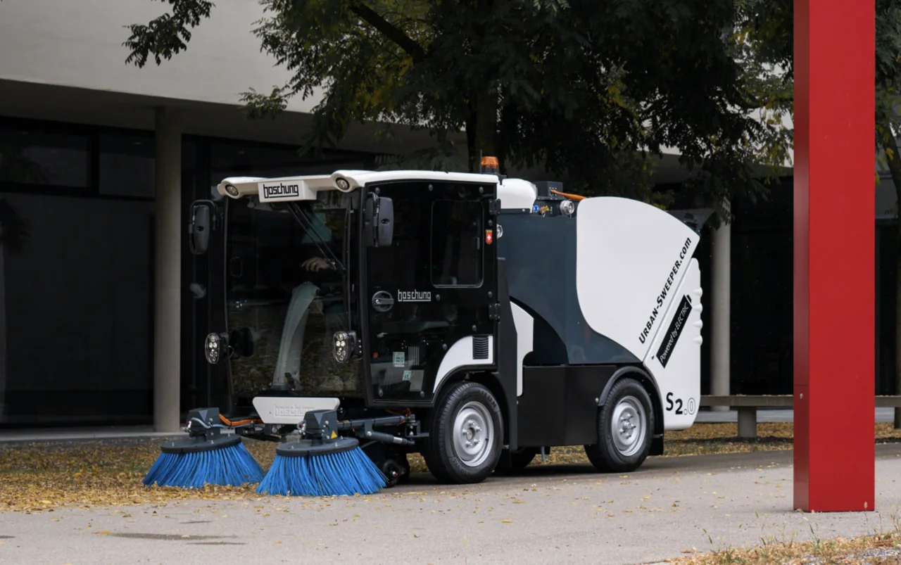 A compact road sweeper has two blue brushes near the ground.