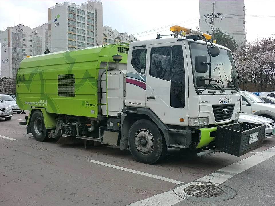 A green-and-white road sweeper shows its cab, side brushes and swept-waste body.