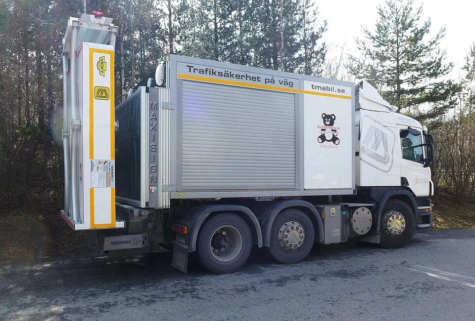 A white roadworks truck carries a tall yellow-and-grey panel at the back.