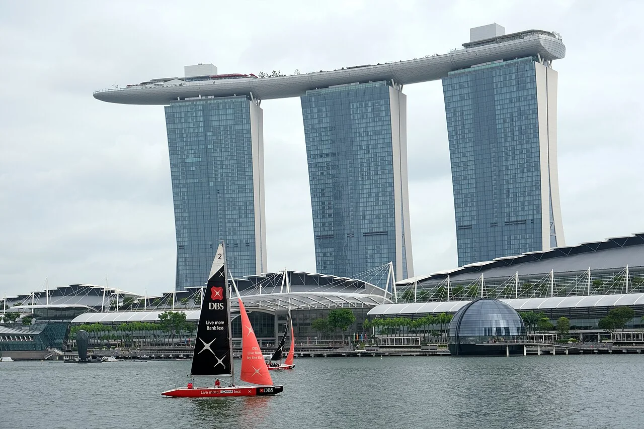 A small sailing boat has a tall dark sail beside Marina Bay Sands.