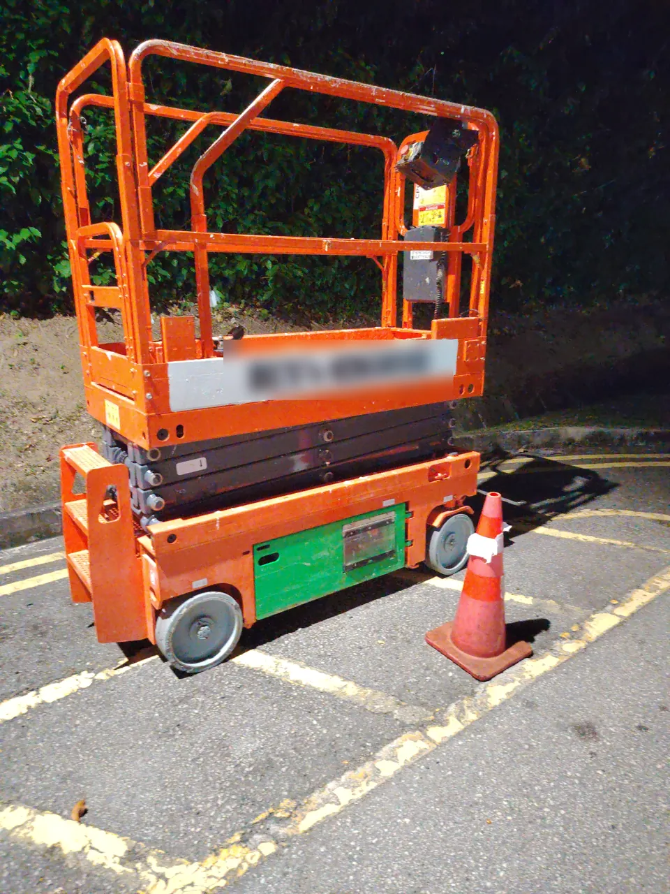 An orange scissor lift with raised guard rails is parked beside a traffic cone at night.