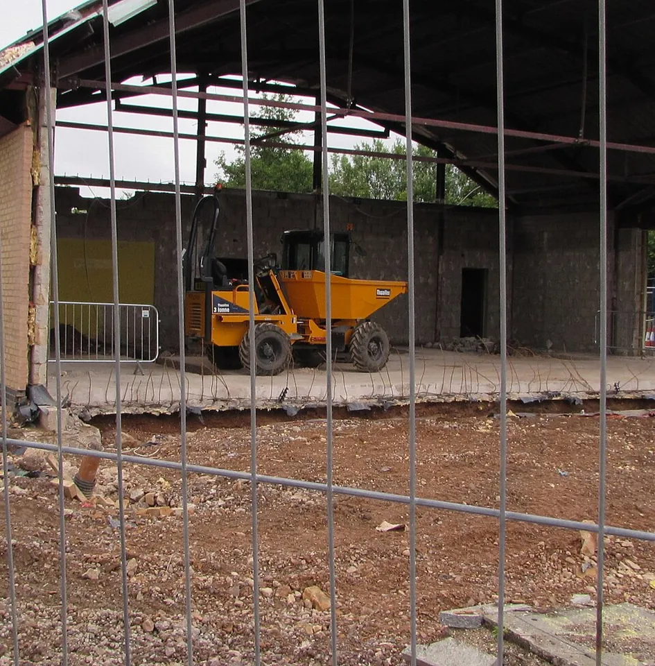 A yellow site dumper stands inside an unfinished building behind a wire fence.