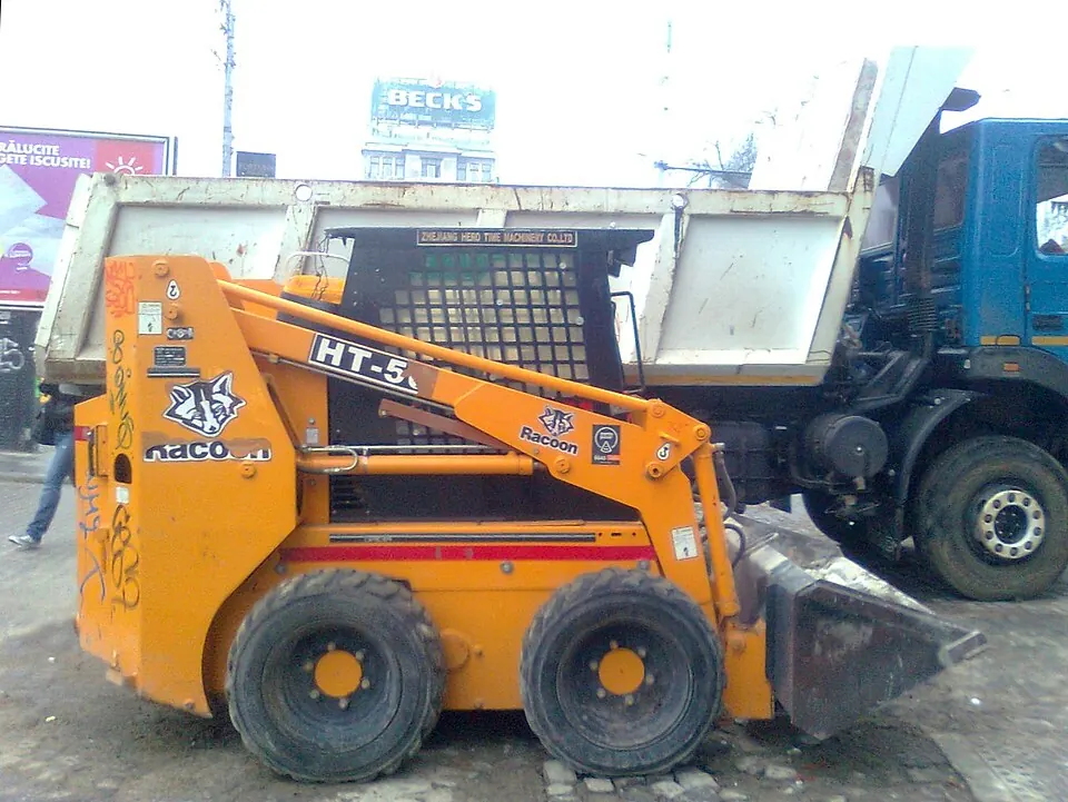 A small orange loader has lifting arms along the sides of its dark cab.