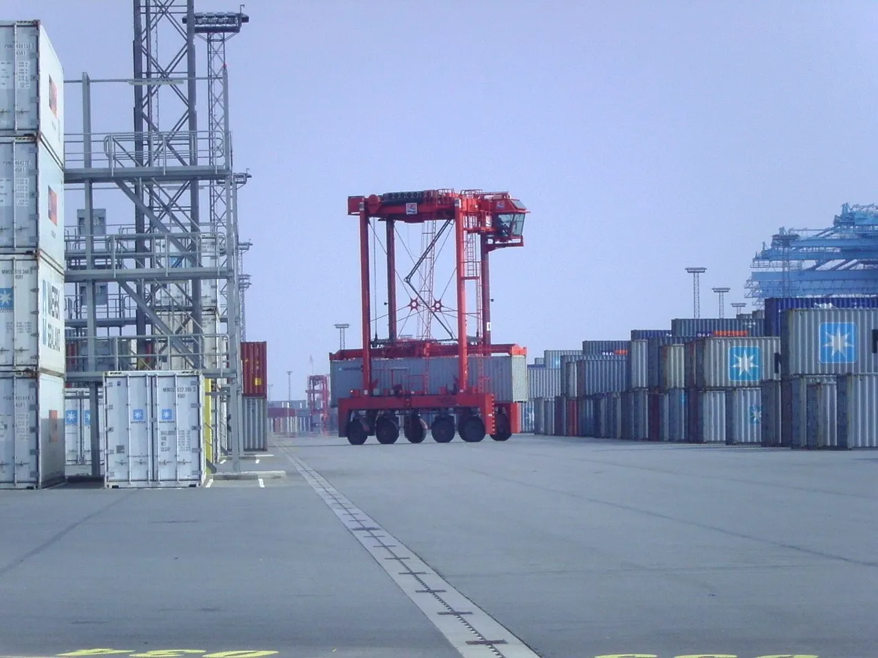 A tall red straddle carrier stands over a lane between container stacks.