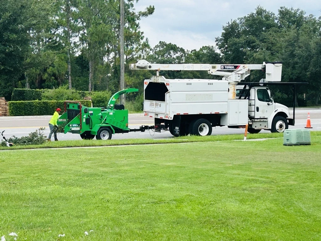 A white bucket truck has its long arm folded above the body, with a green wood chipper behind it.