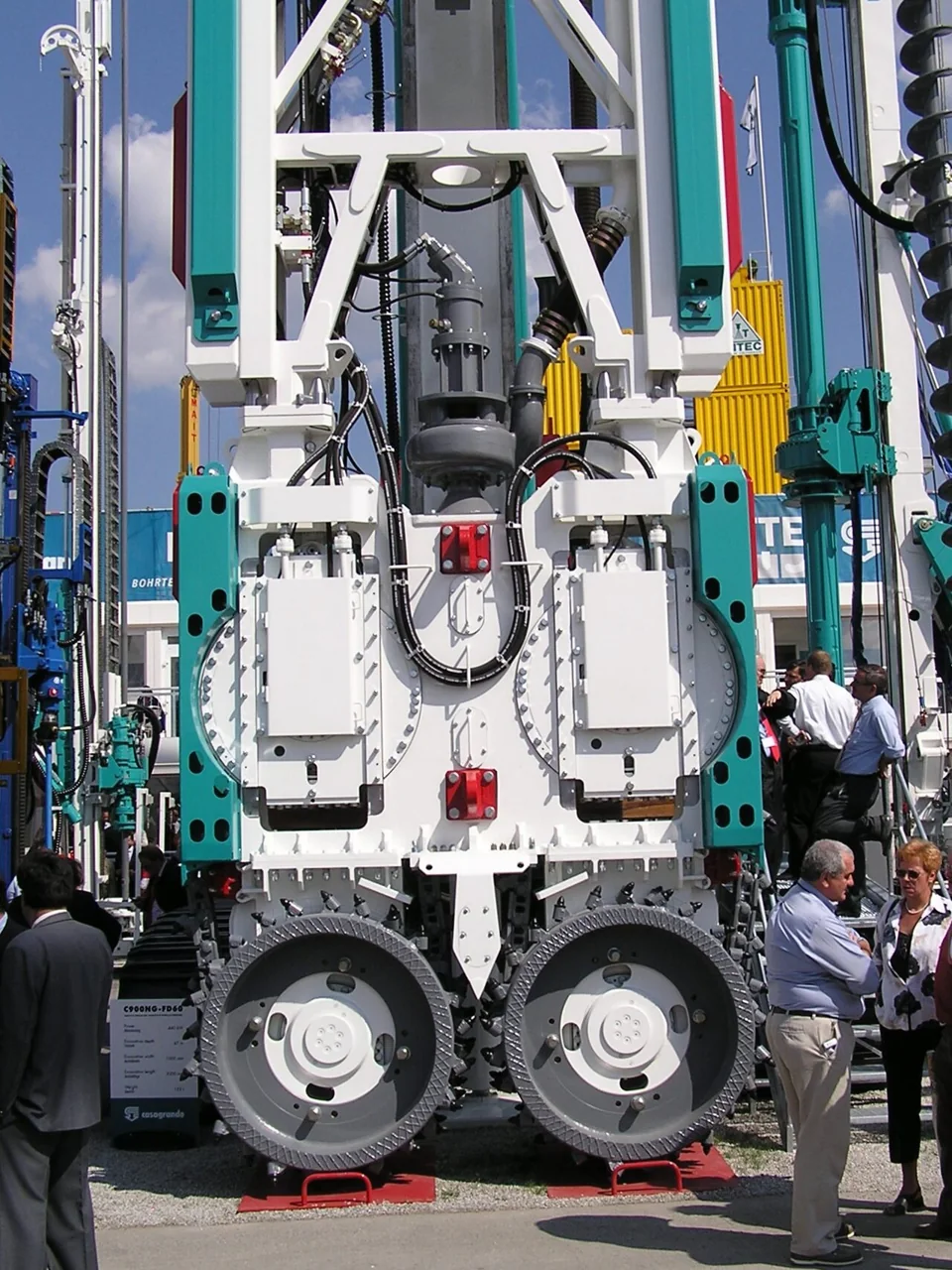 A white and teal trench cutter with two toothed cutting wheels mounted on a mast at an equipment exhibition.