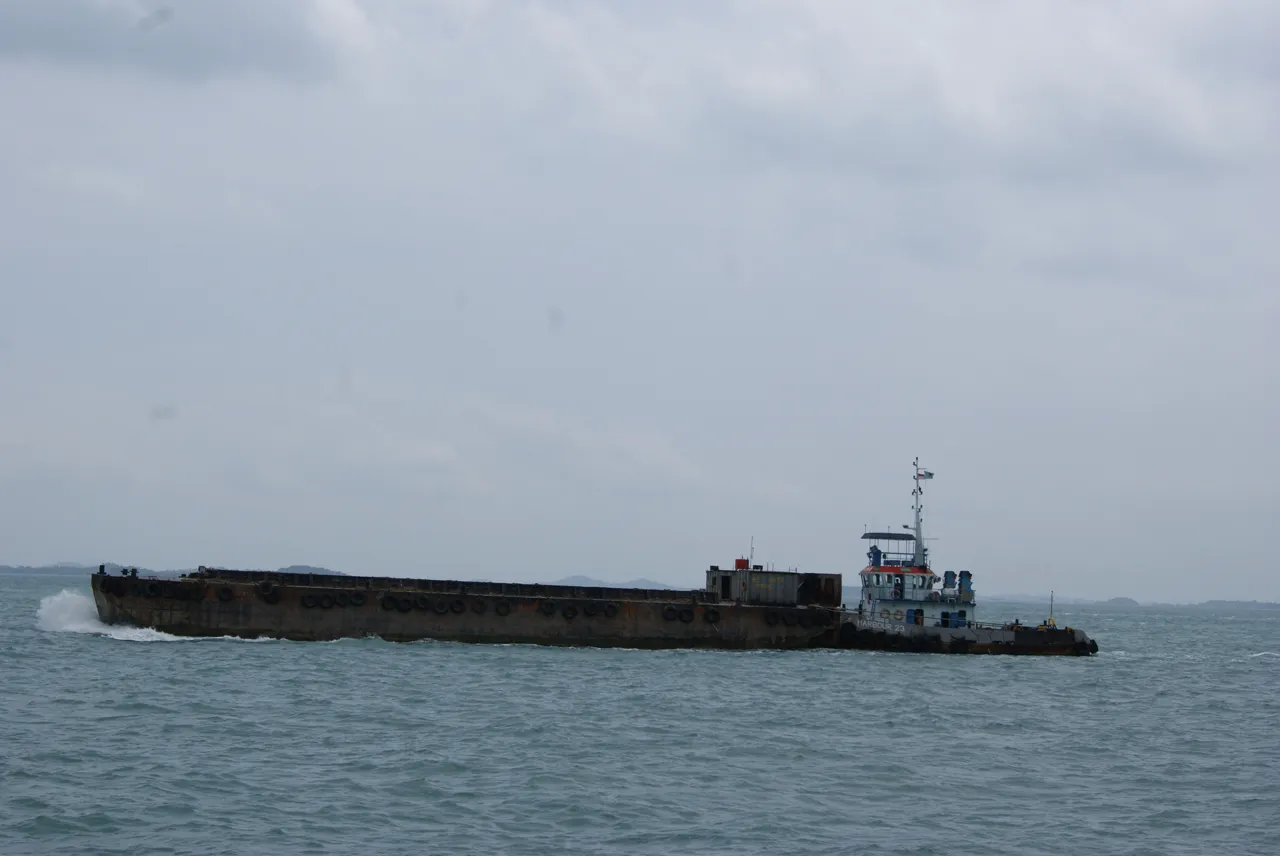 A small tugboat travels beside the end of a long low barge.