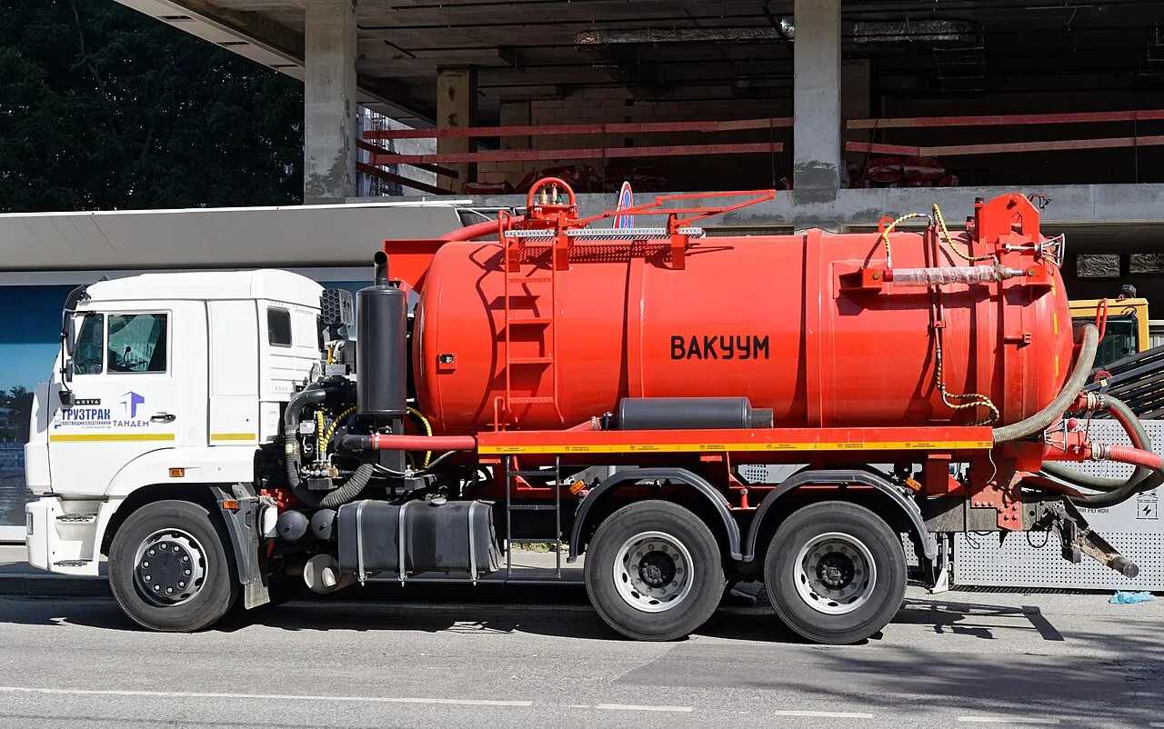 A white truck carries a red vacuum tank with hoses along its side.
