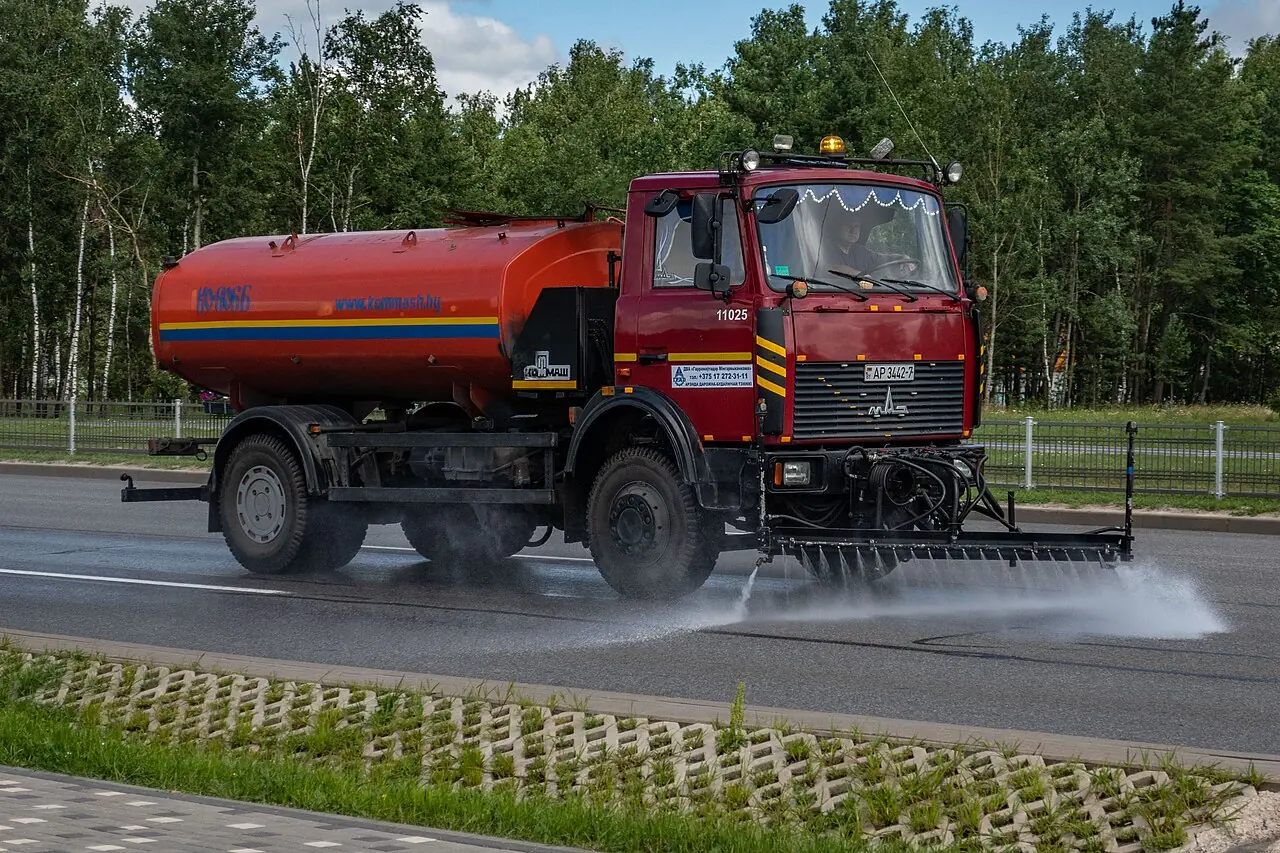 A red water truck sprays water onto the road from a row of front nozzles.