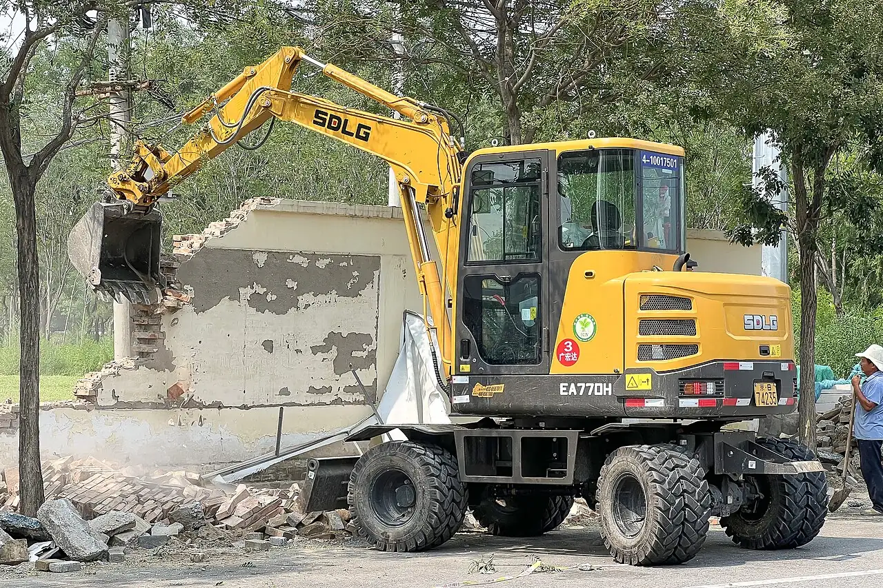 A wheeled excavator uses its bucket beside a broken wall.